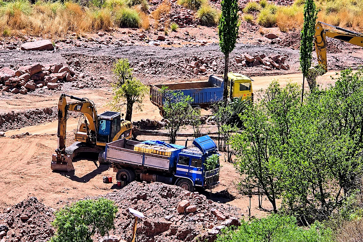 Dos retroexcavadoras y dos volquetas sobre tierra removida y piedras junto al río Tagoni, en Pocoata, Potosí, rodeadas de árboles y laderas secas.